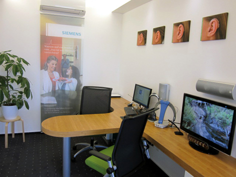 Modernes Hörakustiker-Büro mit Computer, Hörtestgerät und Bildern von Ohren an der Wand.